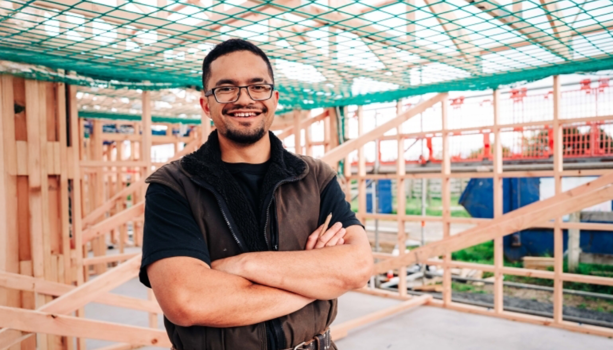 A builder is standing in front of a construction site, smiling