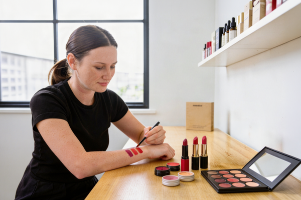A smiling woman does make up color swatches on her arm, comparing lipstick tone