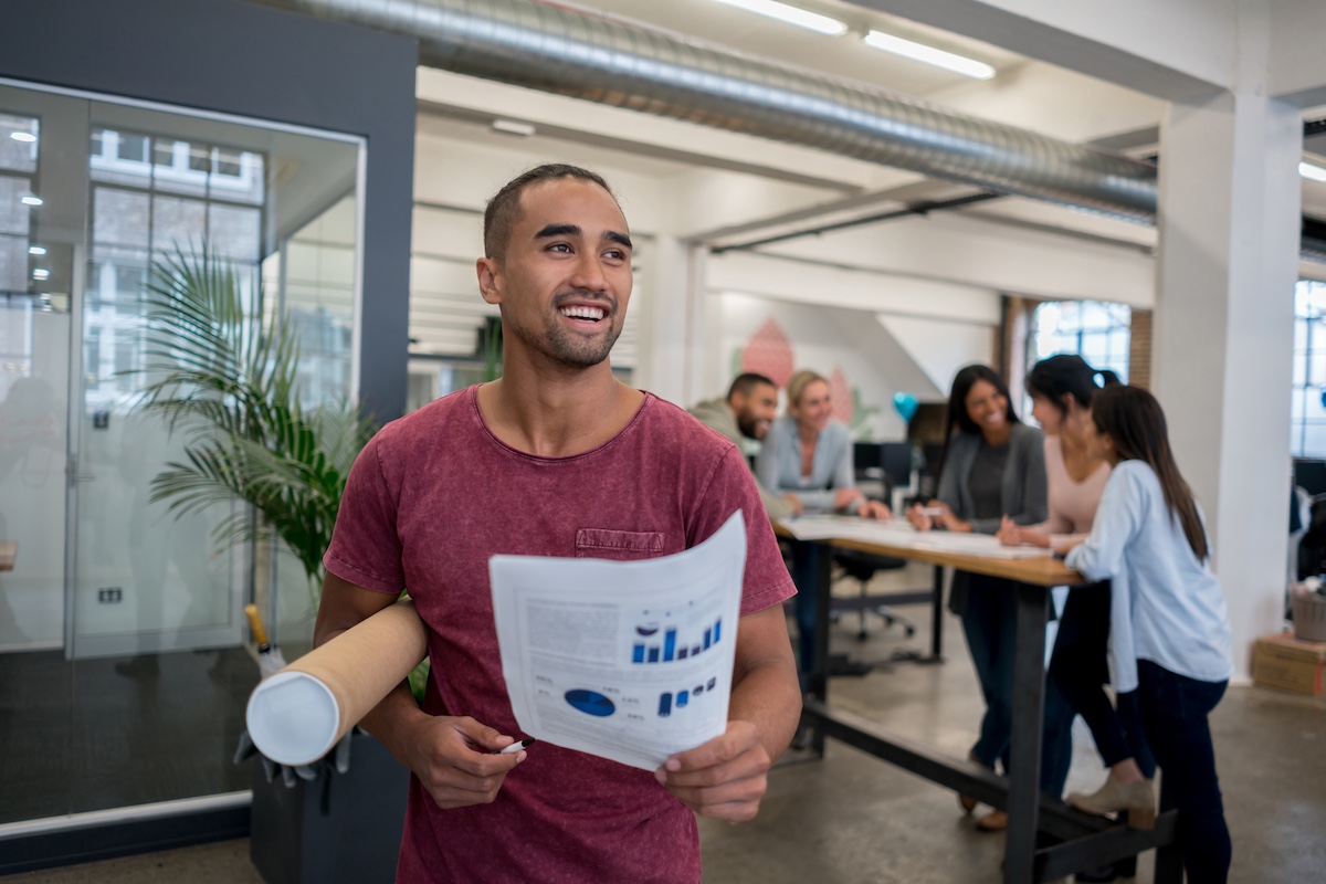 A young man is holding a document with graphs and diagrams, smiling. A mixed group of people on a background are discussing something in an office environment.