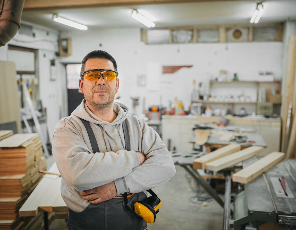 In a workshop, a middle-aged carpetner wearing protective goggles is looking at the camera, smiling