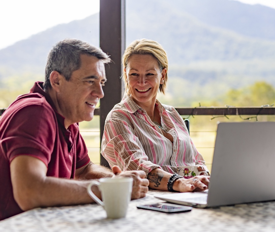 A middle-aged couple are sitting at the table, working on a laptop together, smiling